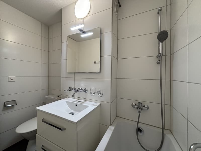 white walls, modern fixtures, white sink, toilet, bathtub, overhead showerhead, illuminated mirror