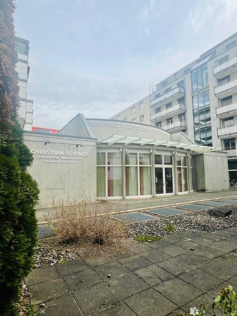 A modern building with gray concrete walls, glass doors and windows, and a small garden in front. The building is labeled as Christian Science. A tall building is on the right side. A bike is parked on the right.
