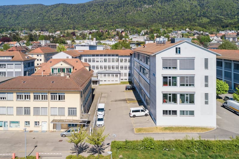 The image shows a view of a town or city, with a mix of residential and commercial buildings. The buildings have a variety of architectural styles, including some with red tile roofs and others with more modern designs. There are several multi-story build