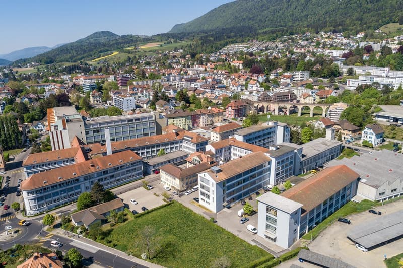 This image shows an aerial view of a city nestled in a mountainous landscape. The city has a mix of modern and traditional architecture, with a combination of high-rise buildings, residential homes, and historic structures. The city is surrounded by lush