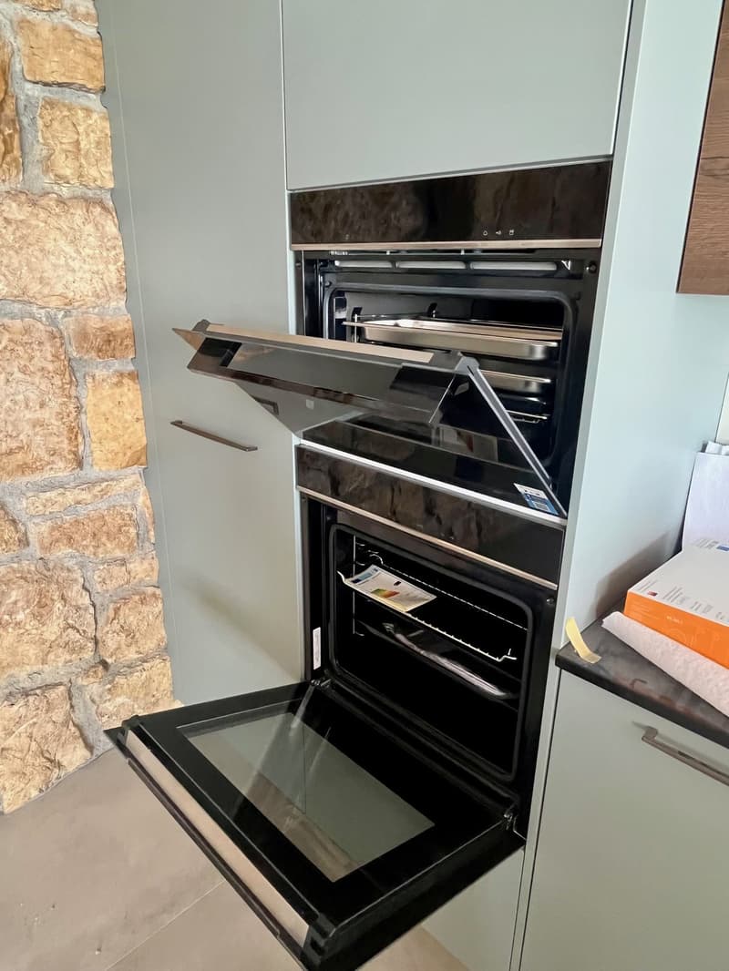 Integrated black oven with a glass door, open, in a kitchen with gray cabinets and a stone wall.