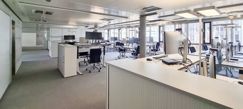 open office layout, white walls, multiple desks, computer monitors, office chairs, ceiling lights