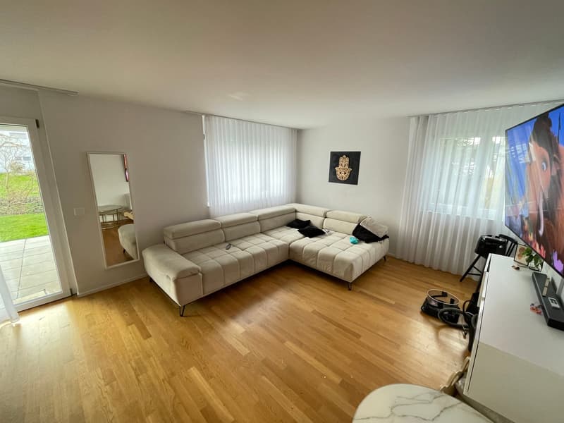 L-shaped sofa, TV, mirror, wooden floor, white walls, white curtains, glass door to balcony