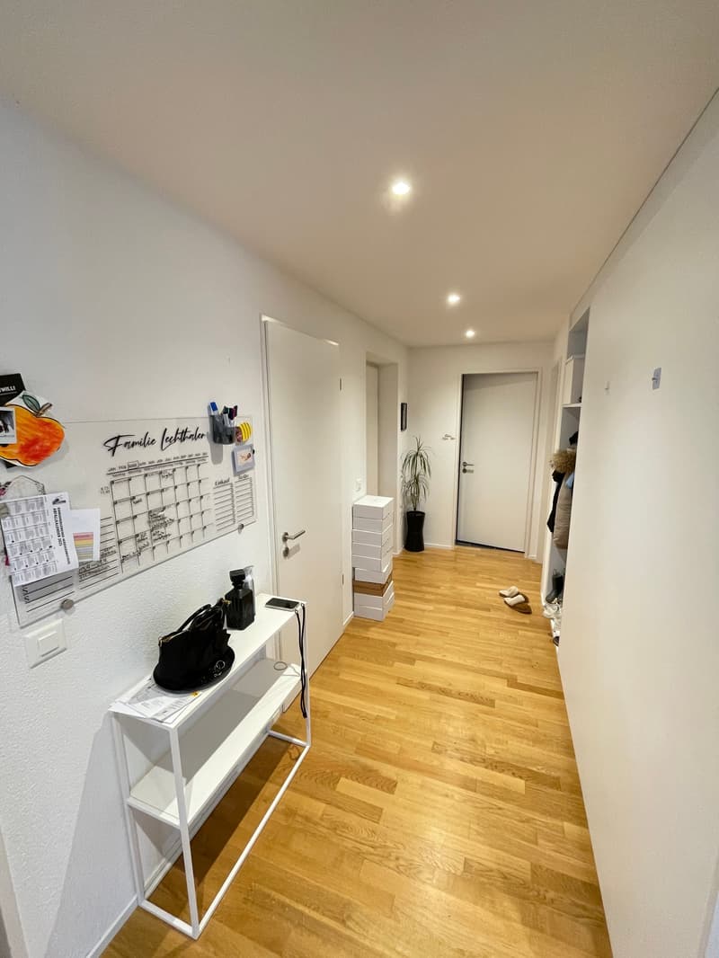 Empty hallway with wooden floor, white walls, white shelf, calendar, potted plant, shoes, storage boxes