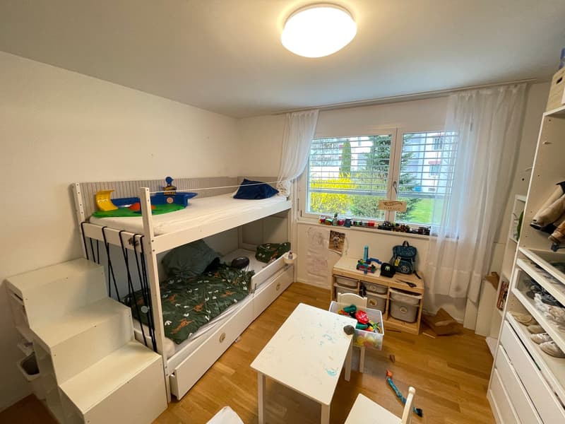 A children's room with a bunk bed, a desk, a chair, and a window with curtains. The room has a wooden floor and white walls.