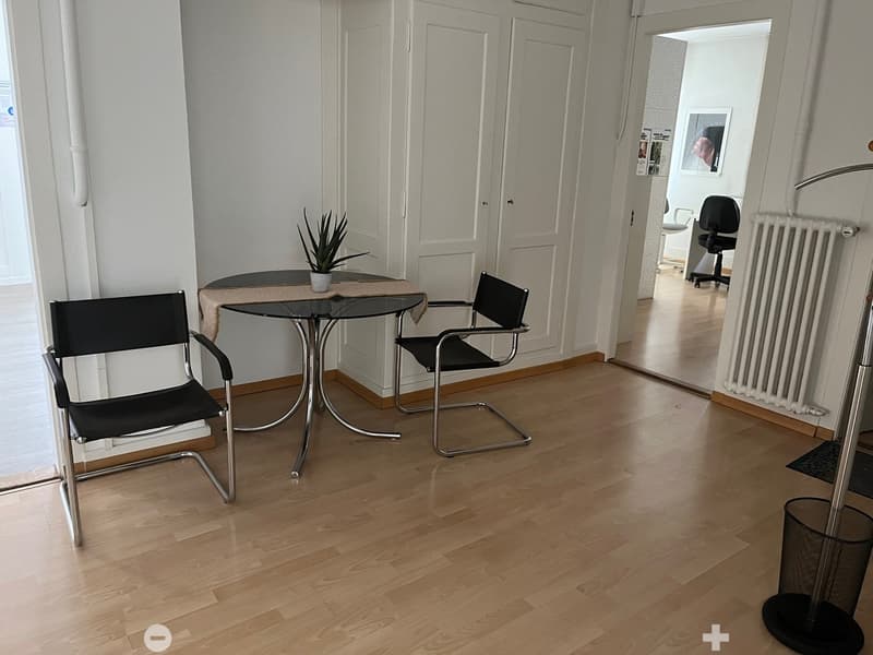 The image shows a small office or dining area with a round glass-top table and two black leather chairs. The room has white walls and hardwood floors, and there is a plant and a framed picture on the wall. The room appears to be part of a larger apartment