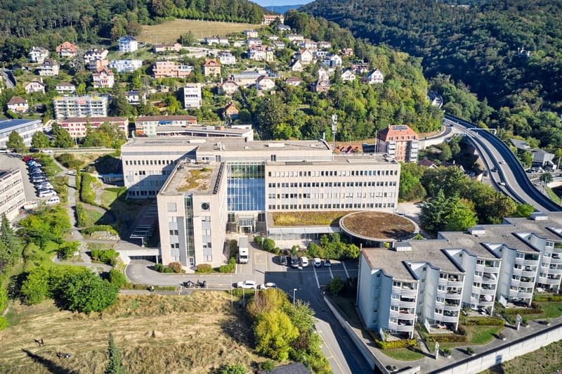 Large office building with green rooftop, several residential buildings, street with parked cars, surrounding nature and a highway