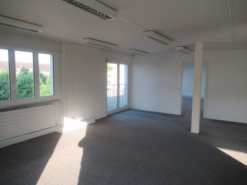 Empty room with carpeted floor, white walls, ceiling lights, double glass windows, sliding glass doors leading to a balcony, radiator, pillar