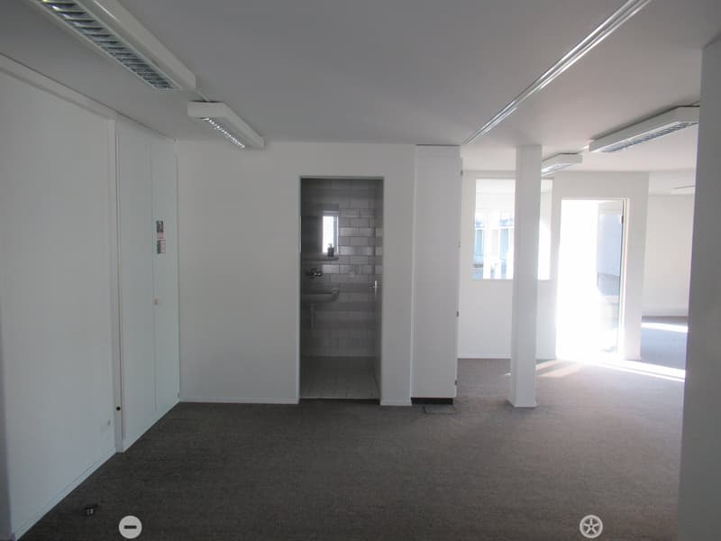 empty room with carpeted floor, white walls, white ceiling, two pillars, bathroom with sink, small window, door leading to another room