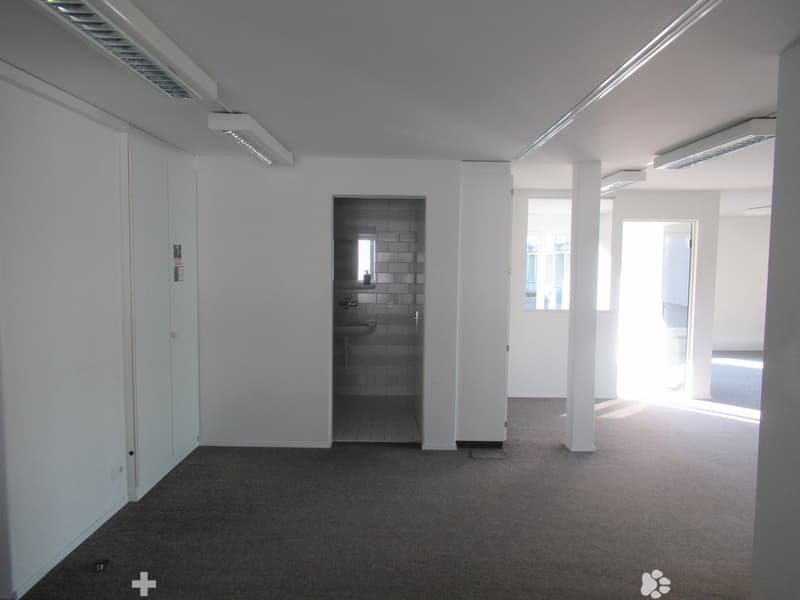 empty room with carpeted floor, white walls, white ceiling, two pillars, bathroom with sink, small window, door leading to another room