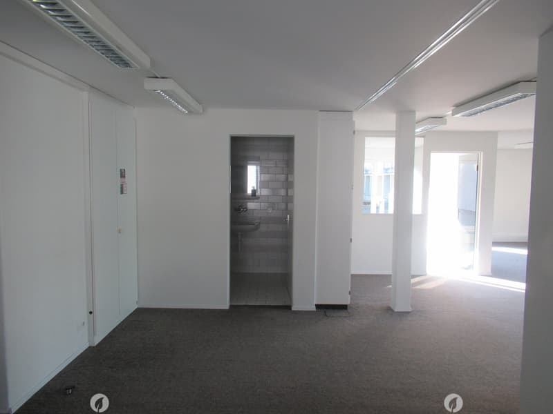 empty room with carpeted floor, white walls, white ceiling, two pillars, bathroom with sink, small window, door leading to another room