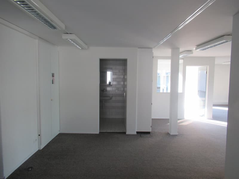 empty room with carpeted floor, white walls, white ceiling, two pillars, bathroom with sink, small window, door leading to another room