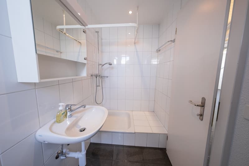 White tiled walls, white tiled floor, small sink, shower with showerhead, mirror, white shower bench