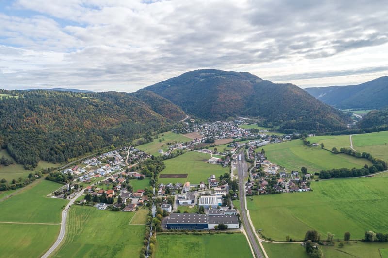 The image shows an aerial view of a small town or village nestled in a mountainous, forested landscape. The town is surrounded by rolling green hills and fields, with a winding road running through the center. There are several buildings and structures vi