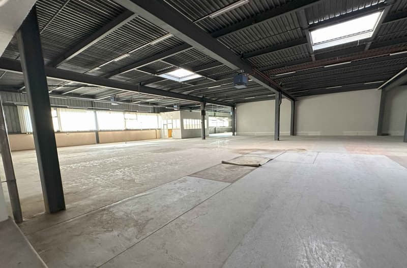 Large open space with concrete flooring, metal ceiling beams, and overhead lighting. The space appears to be a warehouse or industrial facility, with a spacious and unfinished interior.