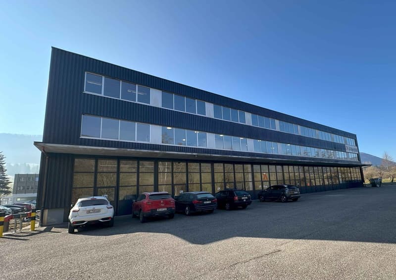 A large, multi-story commercial building with a black metal exterior and numerous windows. There is a parking lot in front of the building with several cars parked.
