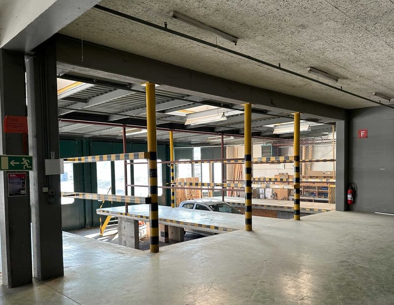 The image shows an indoor parking garage with concrete floors, yellow support columns, and a raised platform for vehicle storage. The space appears to be used for storage or parking, with various shelves and materials visible in the background.