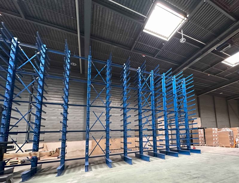 Large industrial warehouse with rows of blue metal shelving units and storage racks. The ceiling is high with skylights, and there are pallets and boxes visible on the floor.