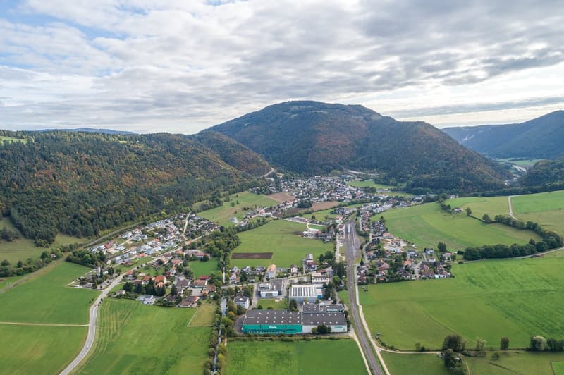 The image shows an aerial view of a small town or village nestled in a valley surrounded by forested hills and mountains. The town has a mix of residential and commercial buildings, with a main road running through the center. The landscape is dominated b