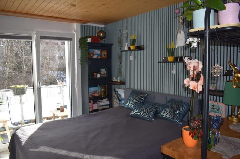 double bed, gray duvet, patterned pillows, wooden side table, balcony view, large window, gray walls, bookshelf, potted plants, decorative items