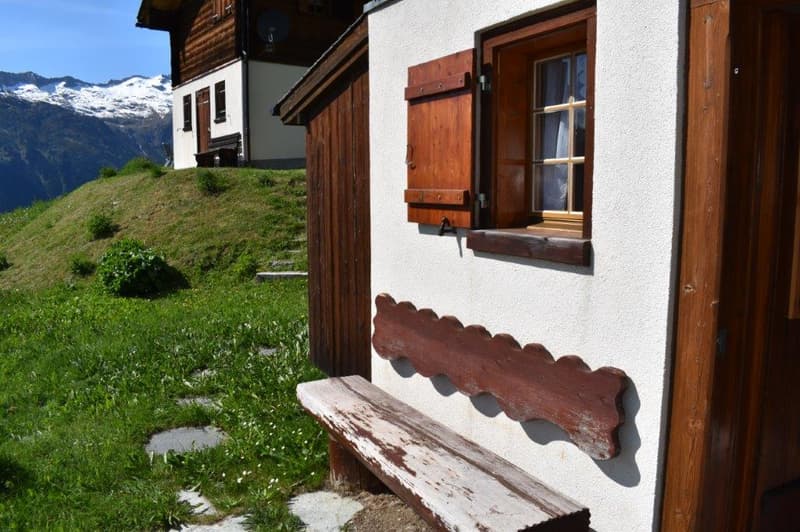 Traditional alpine-style wooden house with white walls, wooden shutters, and a wooden bench outside. The house is situated on a grassy hill with mountains in the background.
