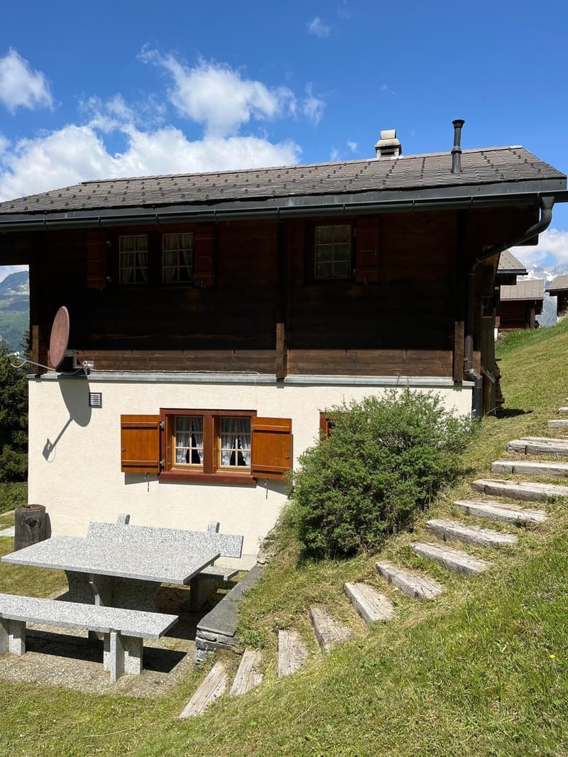 Two-story wooden chalet-style house with a slate roof, surrounded by a grassy area and stone steps leading up to the entrance. The house has large windows with wooden shutters and a balcony on the upper level.