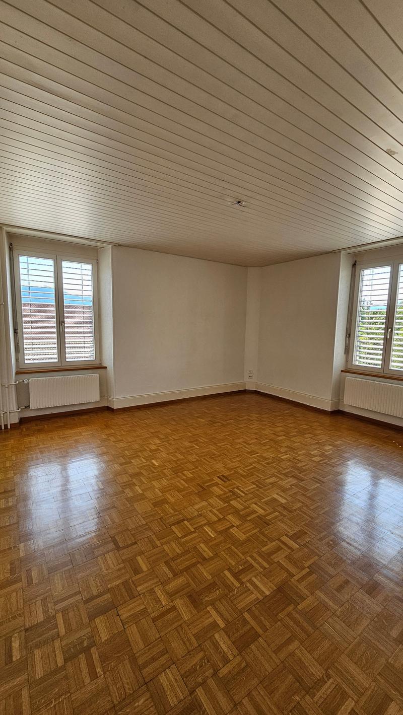 large empty room, wooden parquet flooring, two windows with shutters, radiators, central ceiling beams