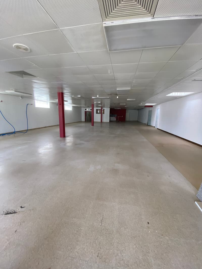 Large open commercial space with concrete flooring, suspended ceiling tiles, and red support columns. The space appears to be unfinished or under renovation.