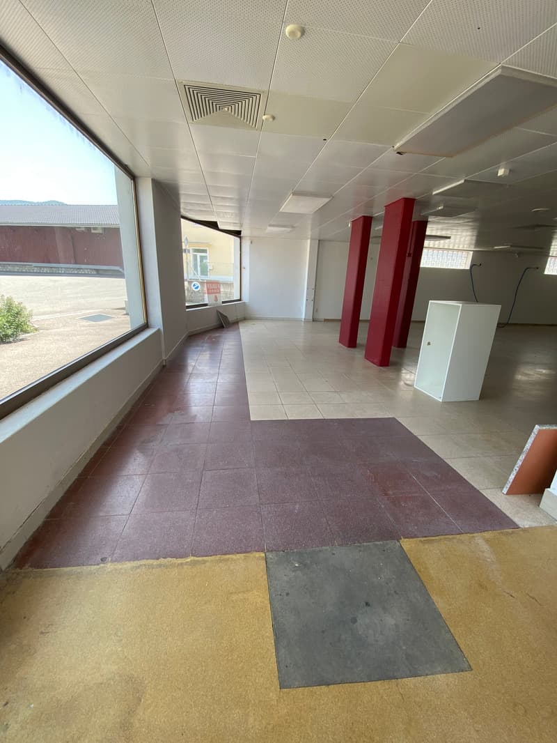 The image shows a large open commercial space with a tiled floor, white walls, and a suspended ceiling with ventilation grilles. There are some red columns visible, and a window overlooking the exterior. The space appears to be an empty commercial or offi