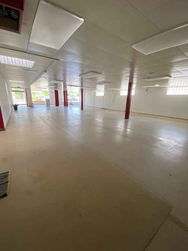 The image shows a large, open commercial space with a concrete floor and white walls. The space appears to be empty, with no furniture or equipment visible. There are several windows along the walls, allowing natural light to enter the space. The ceiling