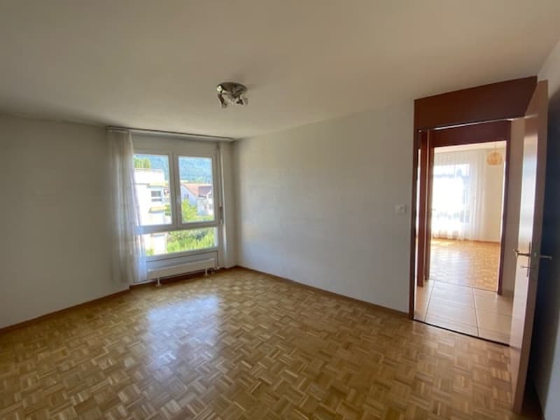 Empty room with white walls, parquet floor, large windows with white curtains, ceiling light, a door leading to another room.