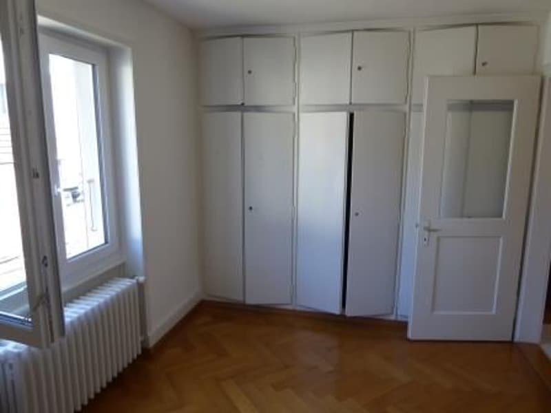 Empty room, white walls, wooden floor, full wall of white wardrobes, large window, white radiator
