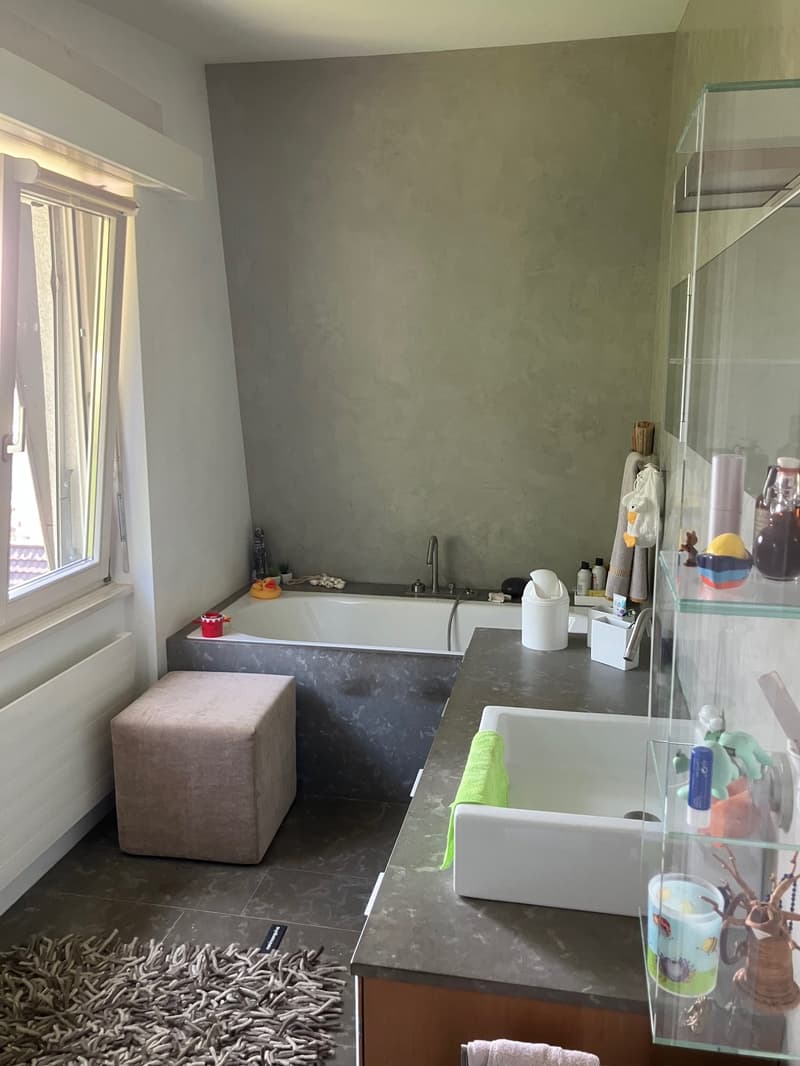 A bathroom with a bathtub, sink, and a shower area. The walls and floor are tiled in gray, and there is a window with a curtain. A fluffy carpet and a small stool are on the floor next to the bathtub.