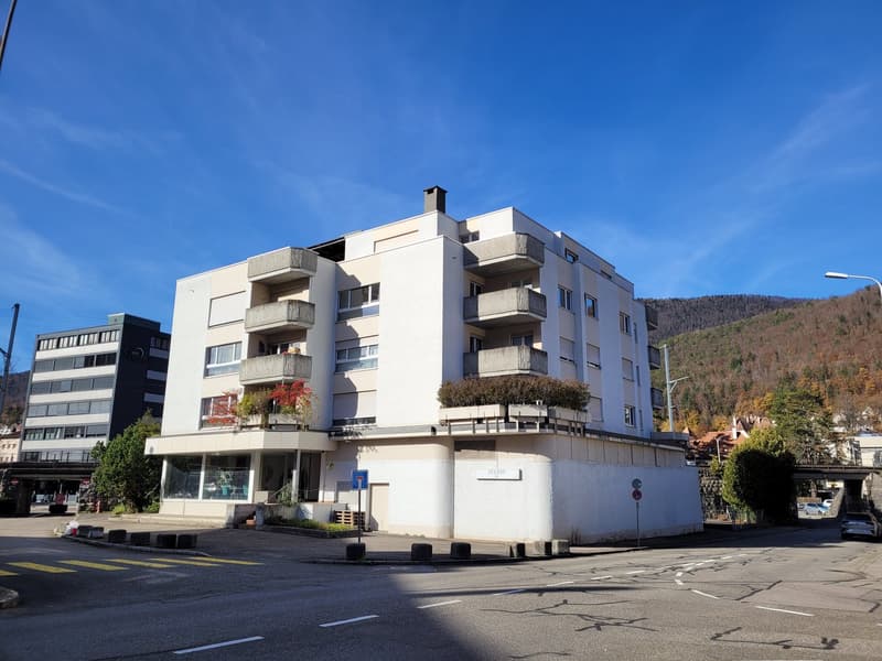A four-story white building with several balconies and greenery on the balconies. A street light and sign is in front of the building.