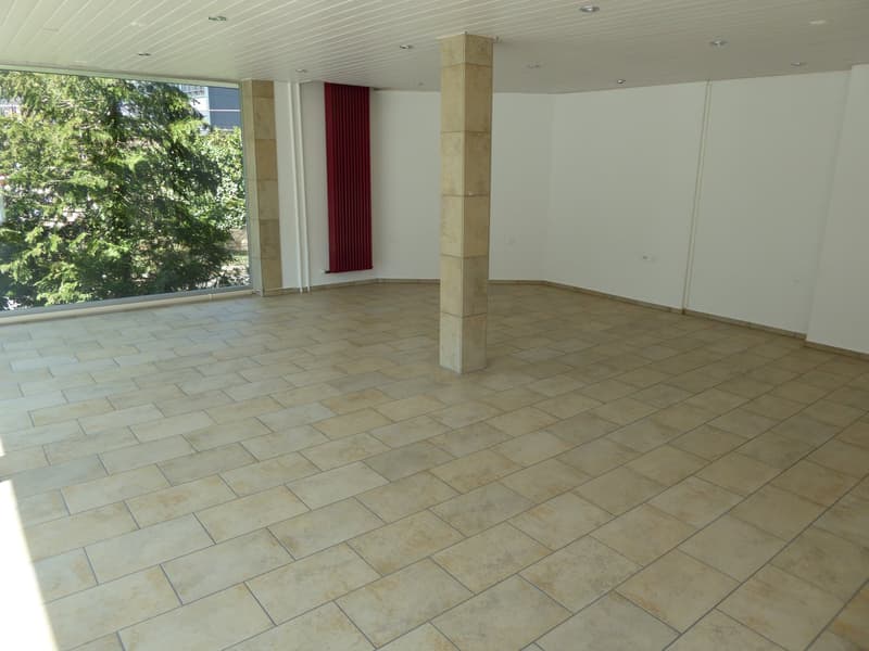 Large, empty room with white walls, tan tiled floor, a central support column, glass window with a red curtain