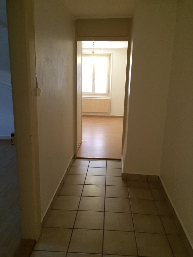 Empty hallway with wooden floors, beige tiles, white walls, window with heater, open door.