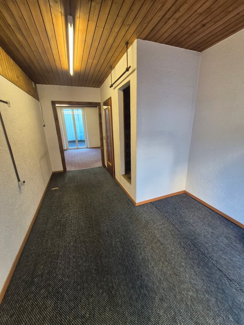 Empty room with wooden ceiling, white walls, gray floor, two doors, one of which leads to another room with a window, and a tube hanging from the ceiling.