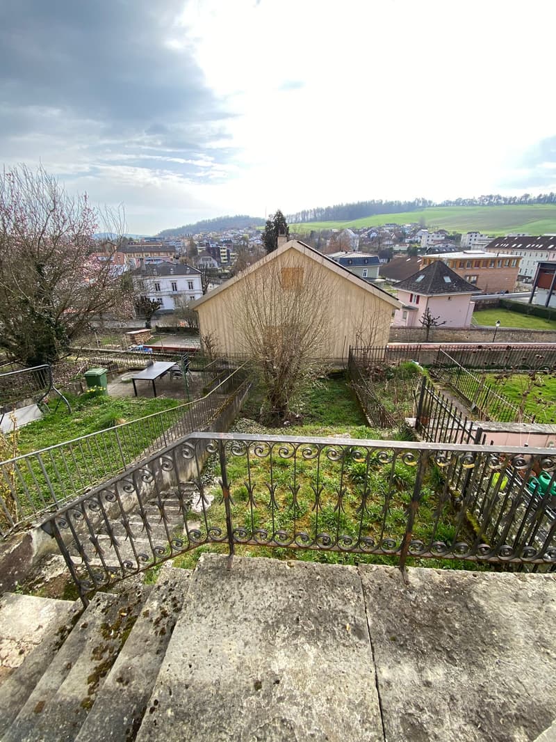 Garden with stairs, metal railing, table, green trash bin, plants, and a view of the town