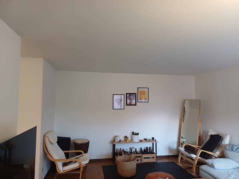 Living room with white walls, wooden floor, a couch, a chair, a table, a mirror, a TV, and a drum.
