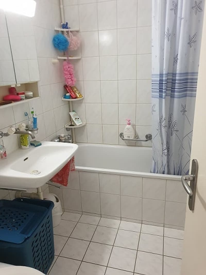 Bathroom with white tiles, white sink, bathtub, shower curtain, shelves with toiletries, and a blue storage basket.