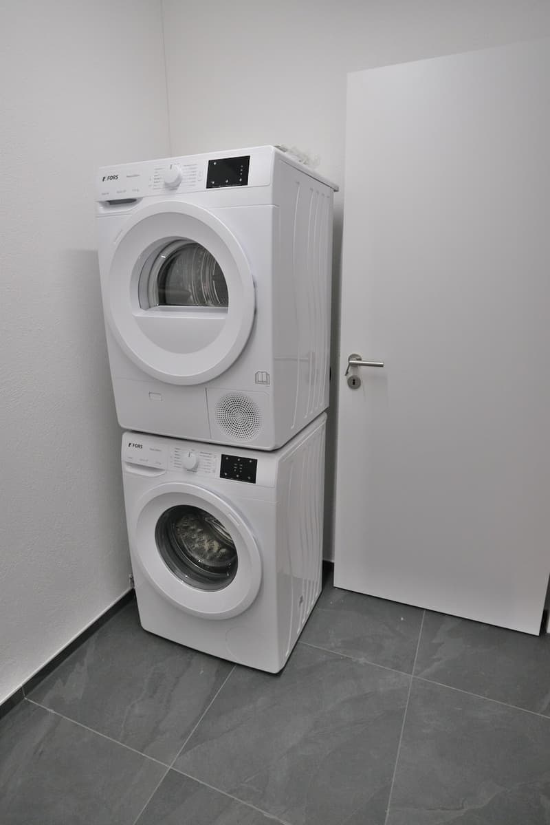A room with a white door, tiled floor, and two stacked white appliances: a washing machine and a dryer.