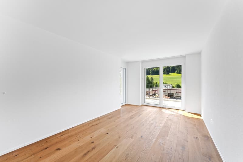 An empty room with wooden floors, white walls, sliding glass doors to balcony, balcony with railings, and a view of a green field