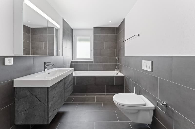 modern bathroom with white bathtub, white sink, white toilet, gray tiles, mirror with light, power outlet