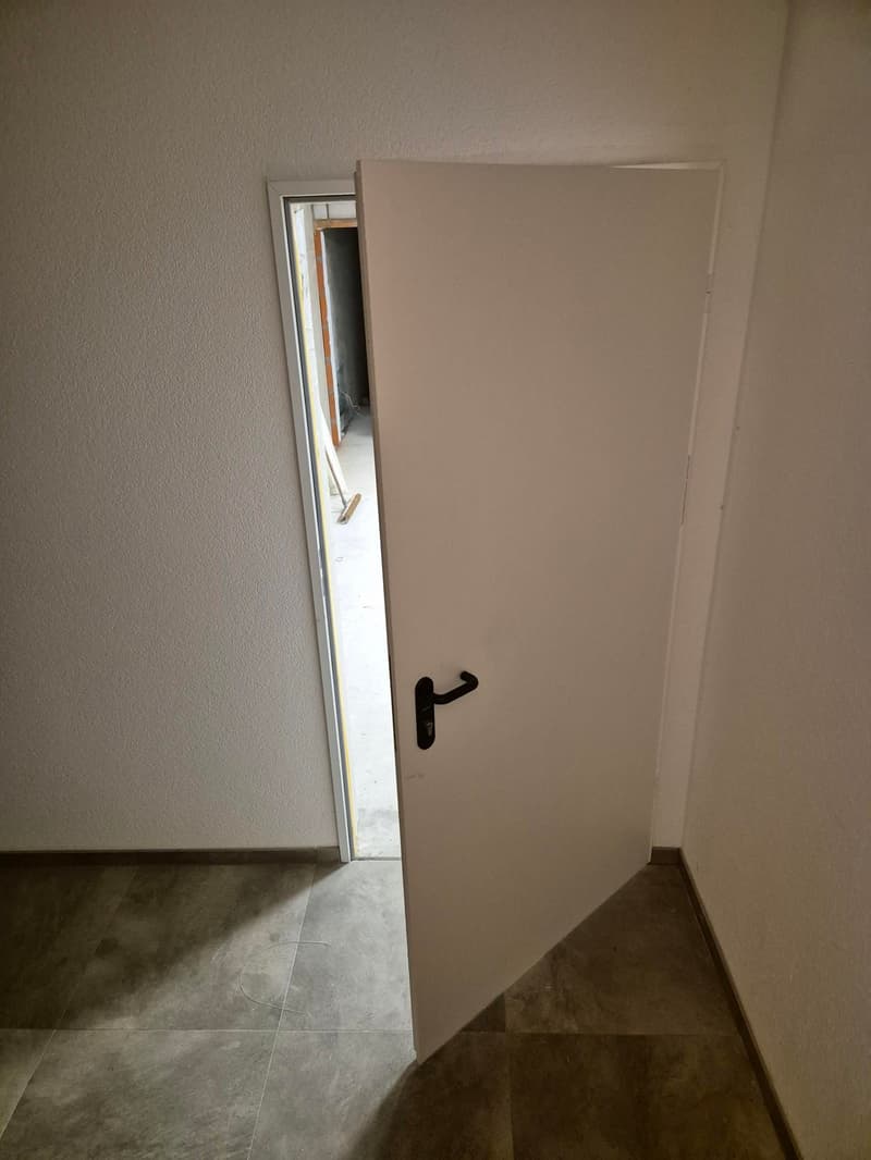 Empty room with tiled floor, open door leading to a partially visible corridor, white walls, black handle on the door