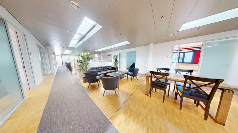 Spacious open-plan office with hardwood floors, high ceilings, and large windows providing natural light. The space includes a lounge area with comfortable sofas and chairs, as well as a dining/meeting area with a wooden table and chairs. The office also