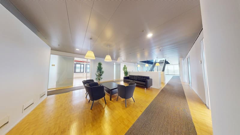 Spacious open-plan office with hardwood floors, white walls, recessed lighting, and a seating area with a sofa, chairs, and a coffee table. There is also a small plant and a staircase visible in the background.
