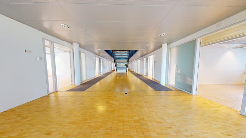 Long, well-lit hallway with white walls, a tiled yellow floor, and multiple doors on both sides. The hallway has a high ceiling with recessed lighting and appears to be part of a larger commercial or institutional building.
