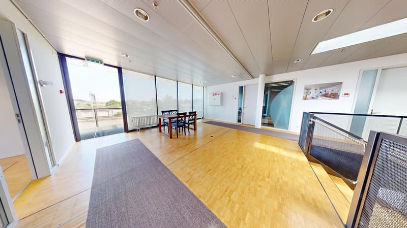 Large open office space with hardwood floors, floor-to-ceiling windows, a dining table with chairs, and a balcony or terrace visible through the windows.