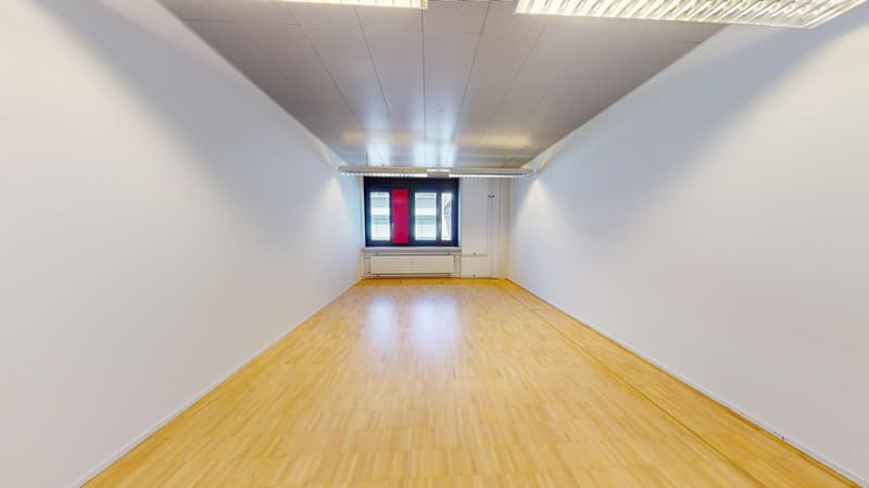 Large open room with hardwood flooring, white walls, and a window with red and blue accents. The room appears to be an empty office or commercial space ready for use.