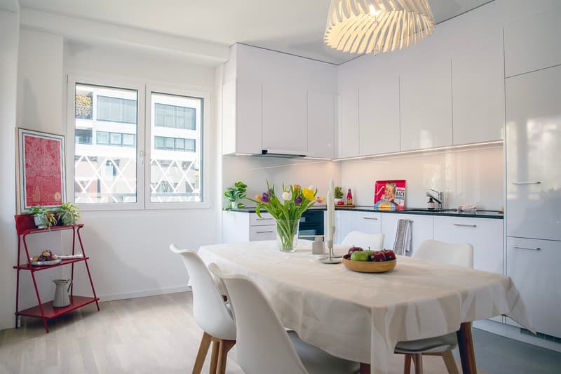Dining room with white walls, wooden floor, white dining table, white chairs, white cabinets, large window, red shelf with plants and pitcher, flowers in vase, bowl of fruit, chandelier